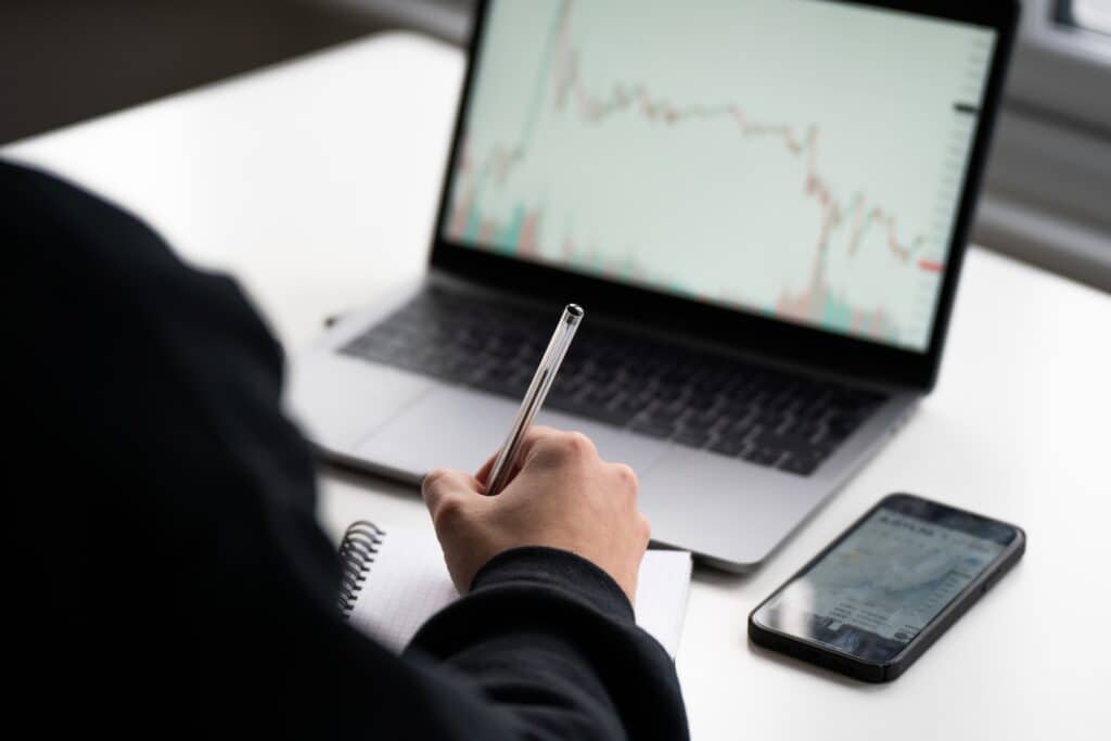 Image of computer, person with pen writing and a cell phone on desk