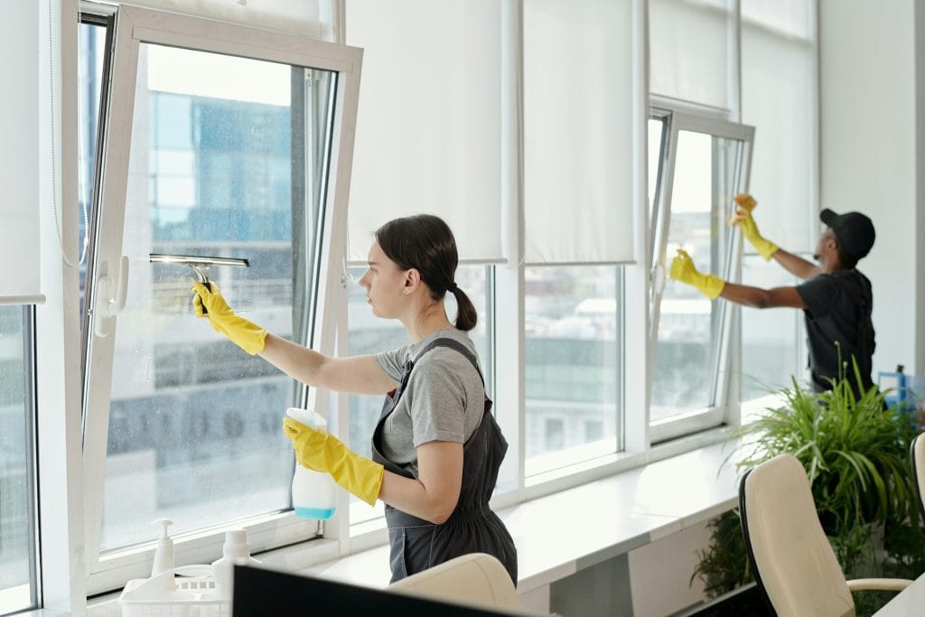 Two cleaning professionals washing large office windows wearing yellow gloves